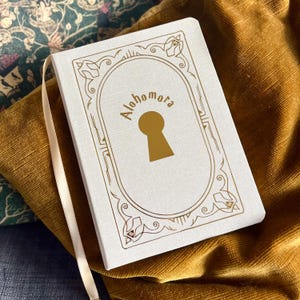 Peut inclure: Un journal en lin blanc avec un motif en relief doré représentant un trou de serrure et le texte "Alohomora".