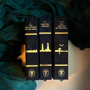 Op de afbeelding: Drie hardcover boeken met zwarte kaften en gouden letters. De titels zijn "The Fellowship of the Ring", "The Two Towers" en "The Return of the King". Elk boek heeft een gouden ontwerp en het symbool van de Ene Ring.