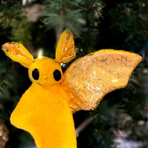 May include: A yellow felt bat ornament with gold glitter wings hanging on a green Christmas tree.