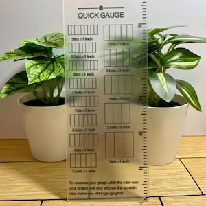 Puede incluir: Una regla Quick Gauge de plástico transparente con marcas y texto en negro, que incluye medidas en pulgadas. La regla está junto a dos pequeñas plantas en maceta con hojas verdes. El texto de la regla dice "Quick Gauge".