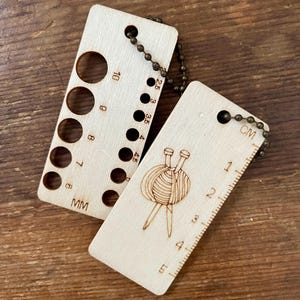 May include: Two wooden knitting tools with attached chain. One tool has numbered holes and measurements in millimetres. The other tool has a centimetre ruler and a graphic of yarn and needles. Both tools are light brown.