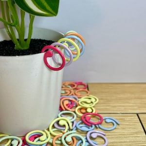 Puede incluir: Una colección de coloridos clips de plantas en espiral en varios tonos pastel, incluyendo rosa, azul, amarillo y naranja. Algunos clips están sujetos a una maceta de cerámica blanca con una planta verde. Los clips están esparcidos sobre una superficie de madera.