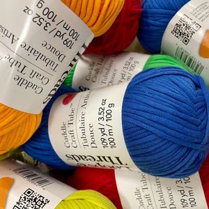 Puede incluir: Primer plano de ovillos de hilo coloridos. La imagen muestra varios rollos de hilo en varios colores, incluyendo amarillo, rojo y azul. Cada rollo tiene una etiqueta que dice "Cuddle Craft Tube" y "109 yd / 100 m / 100 g".