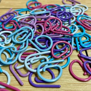 Puede incluir: Una colección de cierres metálicos curvos y coloridos en tonos de azul, morado, rosa y blanco. Los cierres están dispersos sobre una superficie de madera, creando un surtido visualmente atractivo. Estos cierres probablemente se utilizan para manualidades o joyería.