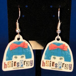 Peut inclure: Une paire de boucles d'oreilles pendantes avec un motif de la com&eacute;die musicale Hairspray. Chaque boucle d'oreille a un fond blanc avec une perruque bleue, un n&oelig;ud rouge et le mot "hairspray" en lettres color&eacute;es. Les boucles ont des crochets argent&eacute;s.