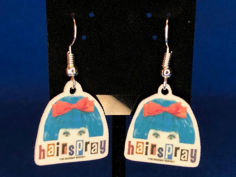 Peut inclure: Une paire de boucles d'oreilles pendantes avec un motif de la com&eacute;die musicale Hairspray. Chaque boucle d'oreille a un fond blanc avec une perruque bleue, un n&oelig;ud rouge et le mot "hairspray" en lettres color&eacute;es. Les boucles ont des crochets argent&eacute;s.
