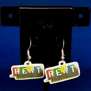 Peut inclure: Une paire de boucles d'oreilles pendantes avec le mot "RENT" en lettres color&eacute;es. Les boucles d'oreilles ont un crochet argent&eacute; et une petite perle transparente. Le fond est un d&eacute;grad&eacute; de bleu.