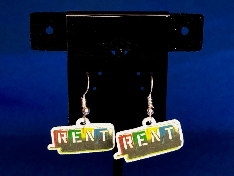 Peut inclure: Une paire de boucles d'oreilles pendantes avec le mot "RENT" en lettres color&eacute;es. Les boucles d'oreilles ont un crochet argent&eacute; et une petite perle transparente. Le fond est un d&eacute;grad&eacute; de bleu.
