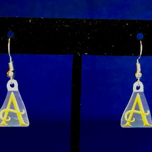 Peut inclure: Boucles d'oreilles en acrylique transparent avec un motif de lettre "A" cursive jaune. Les boucles d'oreilles sont en forme de goutte d'eau avec des crochets d'oreille argent&eacute;s. Le fond est un d&eacute;grad&eacute; de bleu.