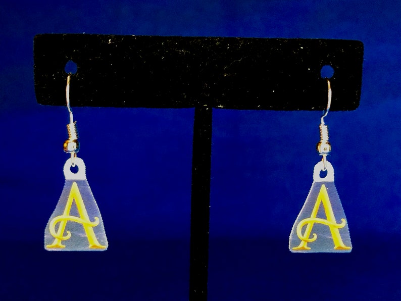 Peut inclure: Boucles d'oreilles en acrylique transparent avec un motif de lettre "A" cursive jaune. Les boucles d'oreilles sont en forme de goutte d'eau avec des crochets d'oreille argent&eacute;s. Le fond est un d&eacute;grad&eacute; de bleu.