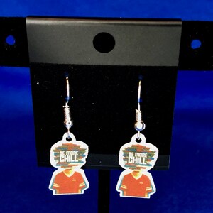 Peut inclure: Une paire de boucles d'oreilles pendantes avec un motif graphique. Chaque boucle d'oreille a un charme blanc en forme de t&ecirc;te avec un motif de chemise rouge et le texte "BE MORE CHILL" dans un style pix&eacute;lis&eacute; color&eacute;. Les boucles d'oreilles sont suspendues &agrave; des crochets argent&eacute;s.
