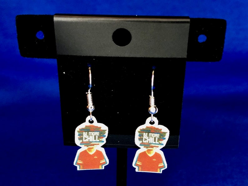 Peut inclure: Une paire de boucles d'oreilles pendantes avec un motif graphique. Chaque boucle d'oreille a un charme blanc en forme de t&ecirc;te avec un motif de chemise rouge et le texte "BE MORE CHILL" dans un style pix&eacute;lis&eacute; color&eacute;. Les boucles d'oreilles sont suspendues &agrave; des crochets argent&eacute;s.
