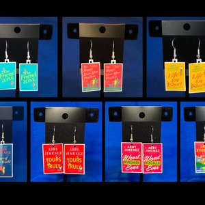 Könnte beinhalten: Ein Set mit 10 Paaren Buchcover-Ohrringen. Die Ohrringe sind silberfarben mit einem rechteckigen Buchcover-Design. Die Buchcover zeigen verschiedene Titel und Autoren. Die Cover sind in verschiedenen Farben, darunter Rot, Blau, Gelb und Grün.