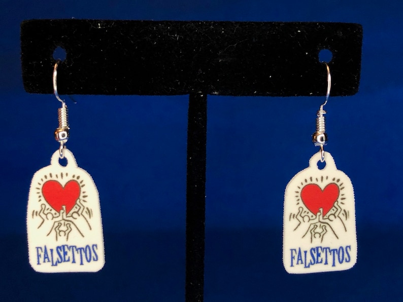Peut inclure: Une paire de boucles d'oreilles blanches avec un motif de c&oelig;ur rouge et le mot "FALSETTOS" en bleu. Les boucles d'oreilles ont des crochets argent&eacute;s et un d&eacute;tail en forme de ressort. Le motif repr&eacute;sente un c&oelig;ur avec des personnages qui tendent la main.