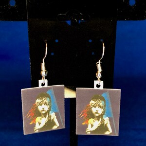 Peut inclure: Une paire de boucles d'oreilles carr&eacute;es avec une impression d'une jeune fille tenant un drapeau, dans les tons de bleu, rouge et blanc. Les boucles d'oreilles ont des crochets d'oreille argent&eacute;s. L'image est sur fond bleu fonc&eacute;.