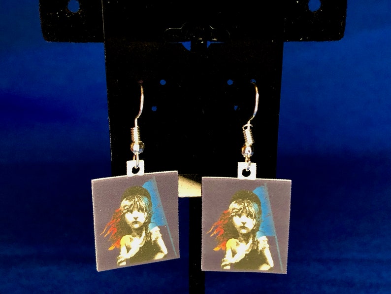 Peut inclure: Une paire de boucles d'oreilles carr&eacute;es avec une impression d'une jeune fille tenant un drapeau, dans les tons de bleu, rouge et blanc. Les boucles d'oreilles ont des crochets d'oreille argent&eacute;s. L'image est sur fond bleu fonc&eacute;.