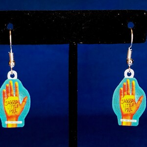 Peut inclure: Boucles d'oreilles pendantes avec un motif de main et le texte "JAGGED LITTLE PILL". Les boucles d'oreilles sont de couleur turquoise et jaune, avec des crochets argent&eacute;s. Un accessoire unique.