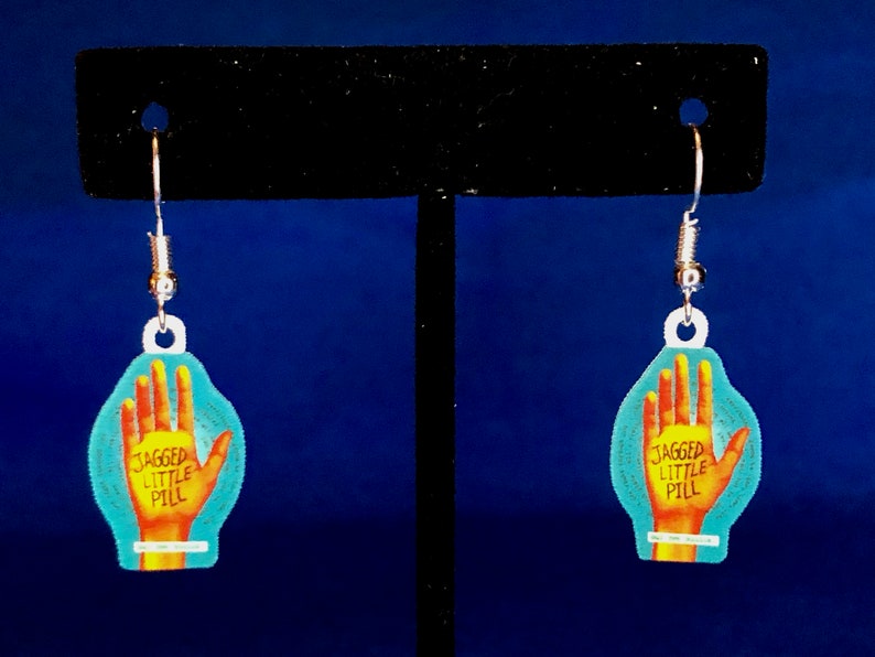 Peut inclure: Boucles d'oreilles pendantes avec un motif de main et le texte "JAGGED LITTLE PILL". Les boucles d'oreilles sont de couleur turquoise et jaune, avec des crochets argent&eacute;s. Un accessoire unique.