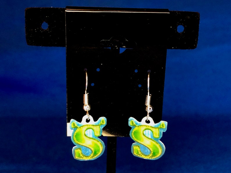 Peut inclure: Une paire de boucles d'oreilles pendantes avec la lettre "S" en vert et jaune, sur fond bleu. Les boucles d'oreilles sont suspendues &agrave; des crochets argent&eacute;s.
