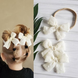 May include: Ivory-colored hair bows and a headband with a matching bow. The bows have a textured, dotted pattern. One image shows the bows in a child's hair, styled in pigtails. The other image shows the bows and headband on a white wooden surface.