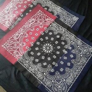 May include: Four bandanas in different colors, red, black, and blue, with a white paisley pattern. The bandanas are folded and stacked on top of each other. The bandanas are made of cotton and are made in the USA.