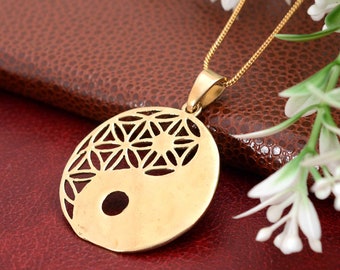 May include: A gold-toned circular pendant necklace featuring a cut-out flower of life pattern on one side and a solid surface with a circular cutout on the other, forming a yin and yang design. The pendant hangs from a thin gold-toned chain.