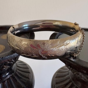 May include: A silver-toned metal hinged bangle bracelet featuring a textured, brushed finish with engraved leaf-like patterns. The bracelet includes a secure clasp mechanism and a thin attached safety chain for added security during wear.