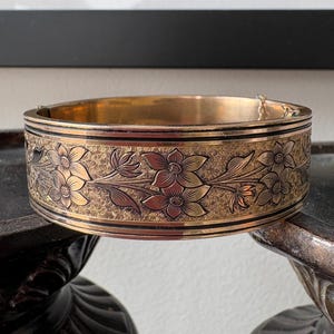 May include: A gold-toned hinged bangle bracelet with an engraved floral design. The bracelet features detailed flowers and leaves, with a textured background. The edges are accented with black lines. The bracelet is displayed on a dark surface.