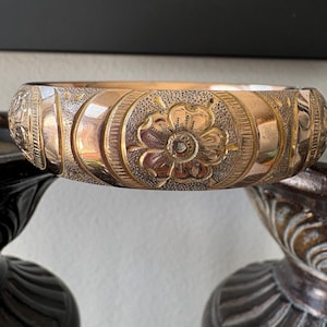 May include: A gold-toned bangle bracelet with intricate floral and geometric patterns. The bracelet features a central flower design and detailed engravings. The metal has a warm, antique appearance, suggesting it may be vintage or vintage-inspired.