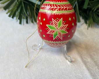 May include: A red, egg-shaped Christmas bauble with green and white star and dot patterns. The bauble has a gold hanging string and is displayed on a clear stand. Festive holiday decoration.