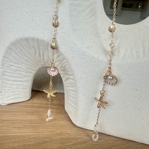 May include: Gold-tone eyeglass chains featuring seashell, starfish, and pearl charms. The chains are delicate and designed to hold eyeglasses. The charms are detailed, adding a coastal aesthetic.