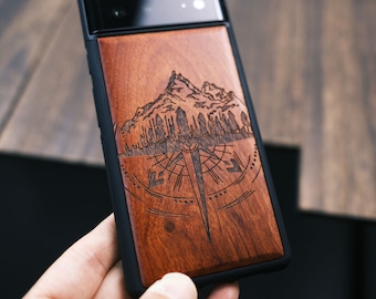 以下が含まれることがあります： コンパスと山のデザインが施された木製携帯電話ケース。ケースは黒色の縁取りのある濃い木で作られています。
