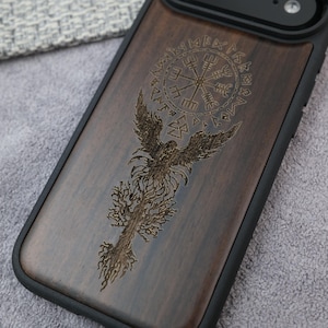 Peut inclure: Coque de téléphone en bois foncé avec un motif gravé. Le motif représente un phénix aux ailes déployées, un symbole circulaire et des motifs complexes. Le boîtier a une bordure noire.