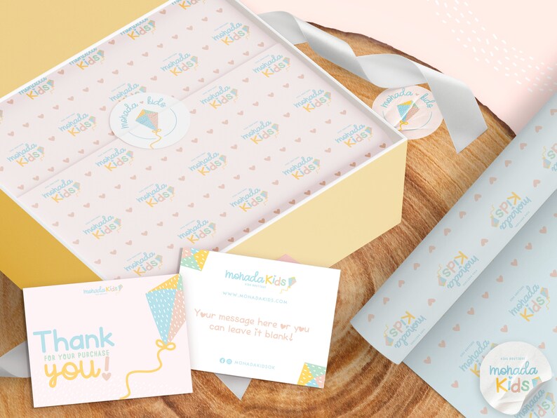 May include: A pink and white gift box with a pink and white patterned tissue paper. The box is filled with small items. The box is sitting on a wooden surface. There is a thank you card with a kite design and the text "Thank you!" on it. There is also a business card with the text "mohada Kids" on it.