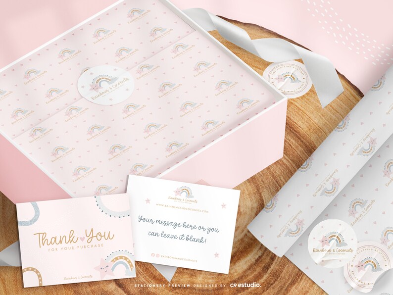 Op de afbeelding: Een lichtroze geschenkdoos bekleed met vloeipapier met een patroon van kleine regenbogen en sterren, vergezeld van twee witte kaarten. E&eacute;n kaart leest Thank You For Your Purchase, en de andere Your message here or you can leave it blank.