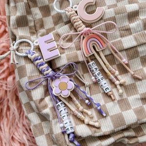 May include: Two handmade keychains with personalized names. One keychain features a purple flower and the name "ELLIE" spelled out in white beads. The other keychain has a rainbow and the name "CASSIE" in white beads. Both have a letter and a braided cord.