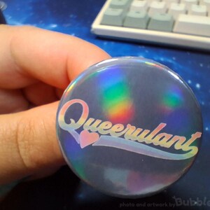 Könnte beinhalten: Ein runder Ansteckbutton mit holografischer Oberfläche, die Regenbogenfarben reflektiert. Das Wort Queerulant ist in einer stilisierten Schreibschrift mittig aufgedruckt, wobei ein kleines rosa Herz den Punkt auf dem i ersetzt.