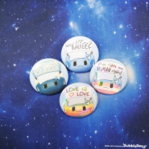 Könnte beinhalten: Vier runde Ansteckbuttons mit Cartoon-Charakteren und Text. Die Buttons haben ein blaues, regenbogenfarbenes und weißes Farbschema. Der Text lautet "Read the Kraken", "Wo Kaffee?", "Love is Love" und "Trans rights are HUMAN rights!"
