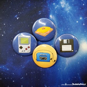 Könnte beinhalten: Vier runde Buttons mit Retro-Designs. Einer zeigt ein pixeliges Sandwich, ein anderer eine klassische Handheld-Konsole, ein dritter eine Diskette und der letzte einen gelben Button mit einem blauen Kassettenrekorder und Kopfhörern.