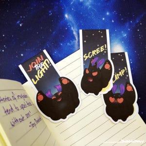 Könnte beinhalten: Drei skurrile Lesezeichen mit einem Cartoon-Fledermaus-Design und den Sätzen "Join the Light!", "Scree!" und "Lamp!". Die Lesezeichen sind schwarz mit roten und lila Akzenten, vor einem Sternenhintergrund.