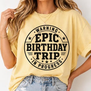 May include: A light yellow t-shirt with a black circular graphic that reads "WARNING EPIC BIRTHDAY TRIP IN PROGRESS." The shirt has short sleeves and is made of a soft material. The model is wearing blue jeans.