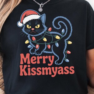 Peut inclure: T-shirt noir avec un chat noir grincheux portant un bonnet de Père Noël et des guirlandes lumineuses. Le texte "Merry Kissmyass" est imprimé en rouge. Fait partie d'une collection sur le thème des fêtes.