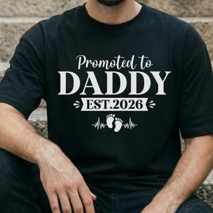 Puede incluir: Camiseta negra con el texto blanco "Promoted to DADDY EST.2026". El diseño incluye huellas de bebé y una línea de latidos. La camiseta de manga corta la lleva una persona sentada en unos escalones.