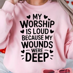 May include: A light pink sweatshirt with the text "MY WORSHIP IS LOUD BECAUSE MY WOUNDS WERE DEEP" in black. The sweatshirt has a crew neck and long sleeves. The image also includes a heart-shaped lollipop.