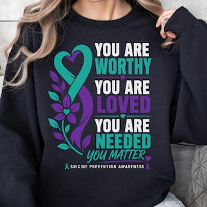 Puede incluir: Una sudadera negra de manga larga con un lazo de concienciación turquesa y morado en forma de corazón con detalles florales. El texto dice: YOU ARE WORTHY, YOU ARE LOVED, YOU ARE NEEDED, YOU MATTER y SUICIDE PREVENTION AWARENESS.