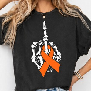 Peut inclure: Un t-shirt noir à manches courtes avec un motif blanc représentant une main squelettique faisant un geste du doigt d'honneur. La main squelettique est entourée d'un ruban de sensibilisation orange vif. Col rond et coupe décontractée.