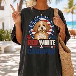 Pode incluir: Uma t-shirt cinzenta escura oversize com o gráfico de um cão Cavapoo castanho e branco dentro de um design circular da bandeira americana. Abaixo do cão, o texto RED WHITE & CAVAPOO aparece em letras vermelhas, brancas e azuis com estrelas.