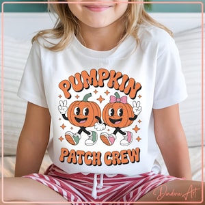 Peut inclure: T-shirt blanc avec un motif rétro. Le texte orange dit "PUMPKIN PATCH CREW". Le graphisme présente deux citrouilles de dessin animé avec des visages, des bras et des jambes, faisant le signe de la paix. Les citrouilles portent des chaussures et sont entourées d'étoiles.