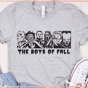 May include: A light grey t-shirt with a black and white graphic of iconic horror movie characters. The text "THE BOYS OF FALL" is printed below. The shirt is shown with white trainers.