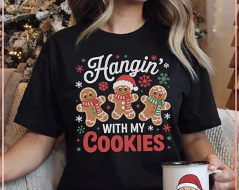 Op de afbeelding: Zwart T-shirt met de tekst "Hangin' with my Cookies" en peperkoekenmannetjes met sjaals en kerstmutsen. Een witte mok met een afbeelding van de kerstman is zichtbaar.